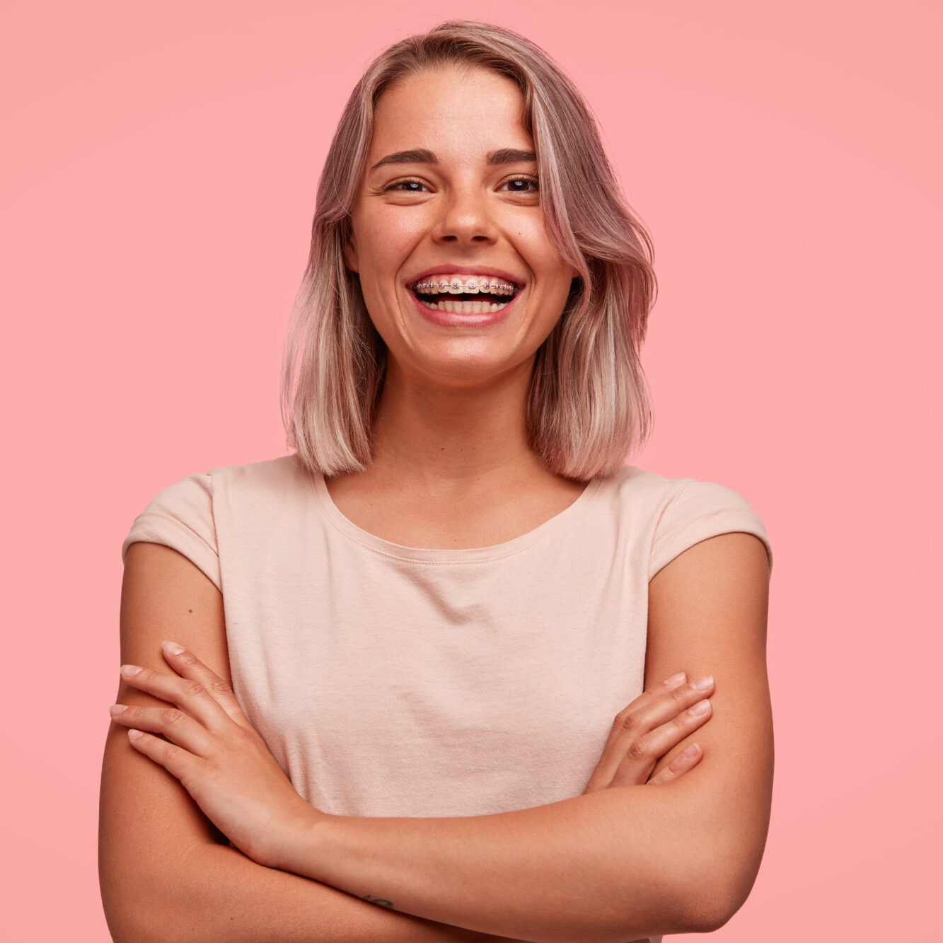 People, happiness and facial expressions concept. Pretty young woman with broad shining smile, keeps hands crossed, being in high spirit, wears braces on teeth, poses alone against pink background